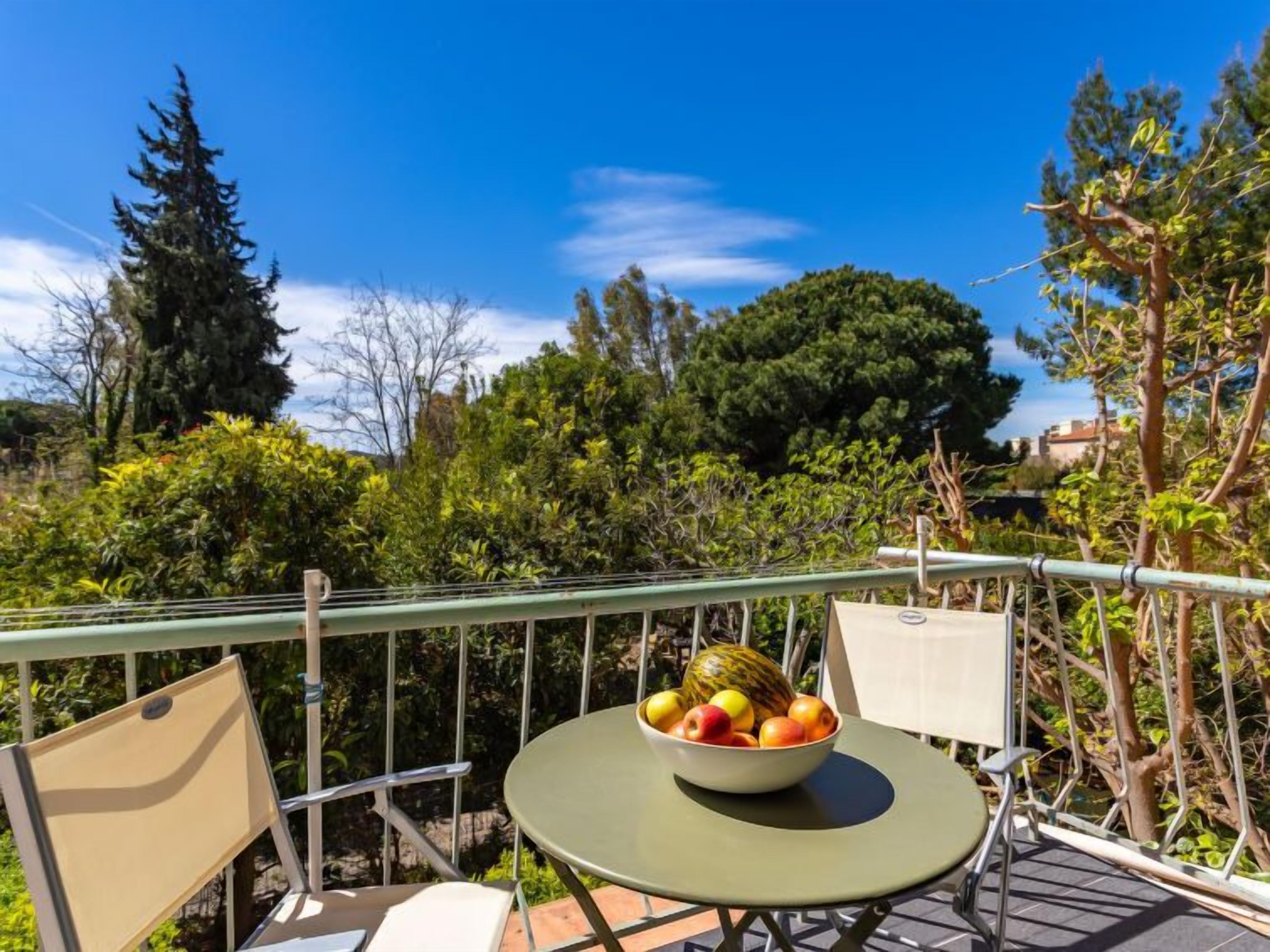 A balcony with a table and chairs, a bowl of fruit, and a view of trees and greenery.