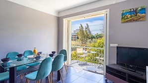 Ein modernes Wohnzimmer mit einem Glastisch, smaragdgrünen Stühlen, einem Flachbildfernseher und einem Balkon mit Blick auf Grünflächen und Gebäude.