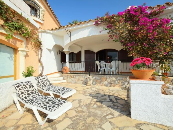 A patio with stone flooring, two lounge chairs, a table with chairs, and a potted plant with pink flowers.