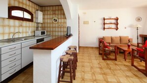 A compact kitchen with a microwave, sink, and a small refrigerator. There is a wooden dining area with a table and chairs, and a sofa. - Peces 3 by Interhome (Peñiscola)