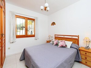 Ein Schlafzimmer mit einem Bett, einem Nachttisch, einer Lampe, einem Fenster mit Vorhängen und einer Wandleuchte.