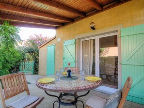 A covered patio with a round table set for dining, green shutters, and a sliding glass door leading to an outdoor area. - Les Marines du Roussillon by Interhome (Saint Cyprien)