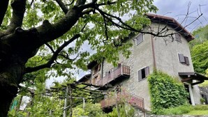 Een tweelaags gebouw van steen met een balkon, omringd door weelderig groen en een boom.