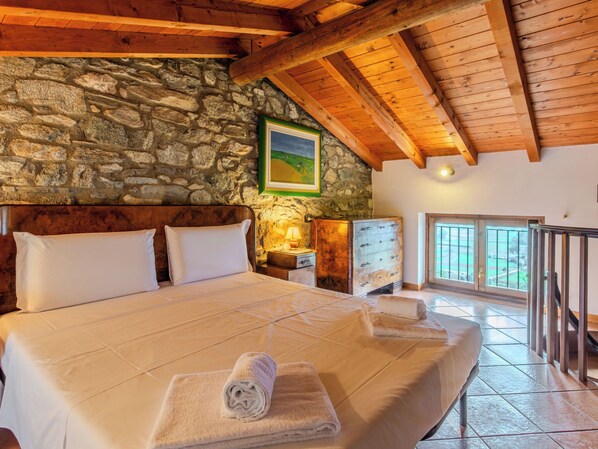 A bedroom with a stone wall, wooden beams, a bed with white linens, a nightstand, a dresser, and a window with a view. - Cà del Fritz by Interhome (Sorico Albonico)