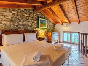A bedroom with a stone wall, wooden beams, a bed with white linens, a nightstand, a dresser, and a window with a view.