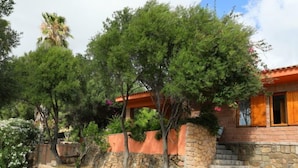 Un bâtiment aux murs de pierre et au toit recouvert de tuiles rouges, entouré d’une végétation luxuriante et d’arbres.