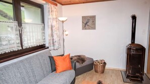 En hyggelig stue med en grå sofa, en orange pude, trægulv, pejs og et vindue med blondegardiner.