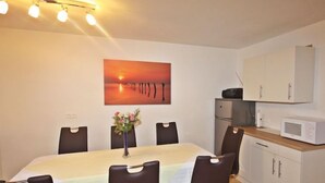 Spiseområde med et bord, stole, en blomstervase, et maleri af en solnedgang, en mikrobølgeovn og et køleskab.