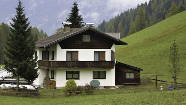 En toetages chalet med balkon, omgivet af en indhegnet grund og med et bjerg i baggrunden.