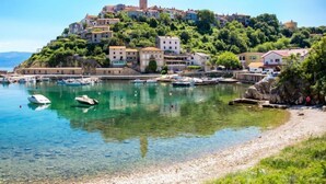 Un pueblo costero con edificios en una colina, un puerto claro con barcos y un litoral rocoso.
