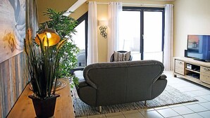 A living room with a sofa, a TV, a plant, and a large window with curtains. - Witthuus-Langeoog by Interhome (Norddeich)