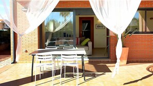 Un patio al aire libre con una mesa y sillas, un ventanal de vidrio que da a un espacio interior y un muro de ladrillo.