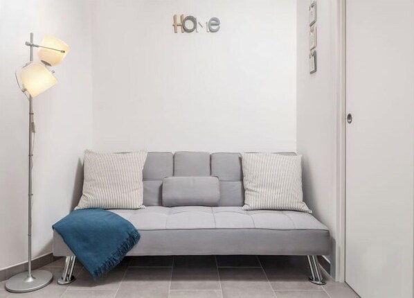Un salon moderne avec un canapé gris, des coussins à rayures et un plaid bleu. Une lampe de sol à deux abat-jour se trouve à gauche.