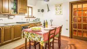 A kitchen with wooden cabinets, a dining table with a checkered tablecloth, and a hanging light fixture. - Tribolina by Interhome (Cavriglia)