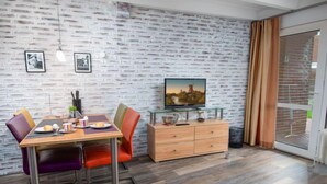 Comedor con pared de ladrillo, mesa de madera y sillas color naranja. Hay un televisor sobre un aparador de vidrio y una ventana con cortinas.