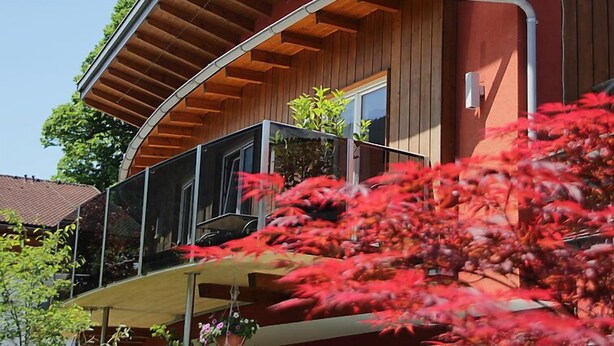 Une maison moderne avec une façade en bois, un balcon doté d’une rambarde en verre et, au premier plan, une plante à fleurs rouges.