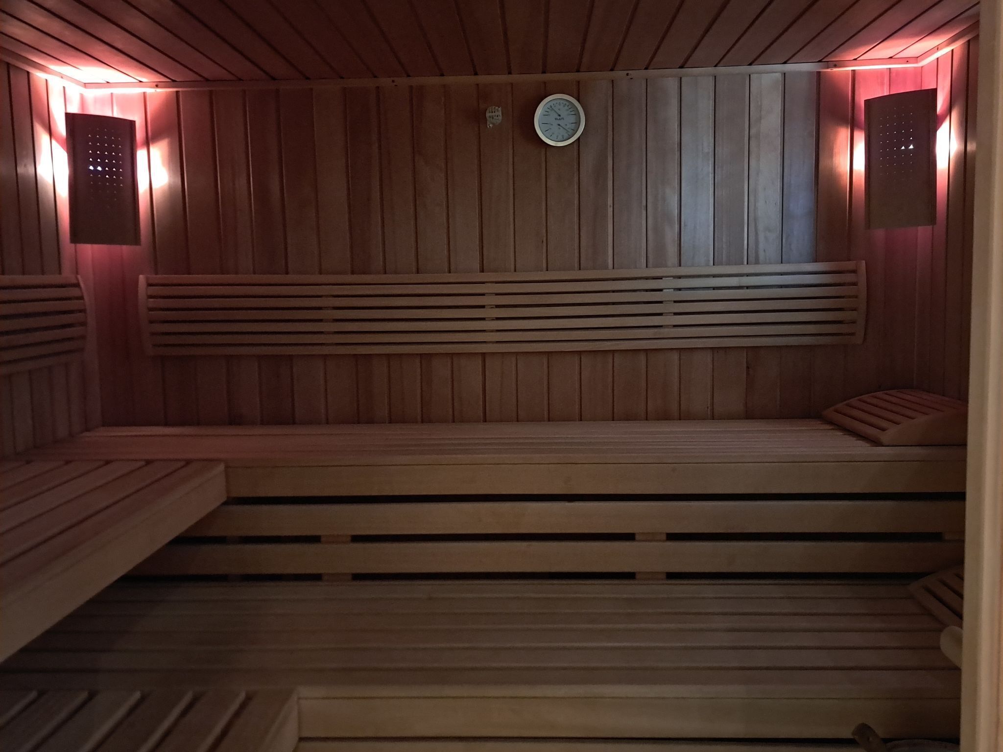 Una sauna con bancos de madera y asientos con listones, un reloj en la pared y luces fijadas a la pared.