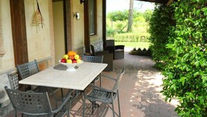 Une terrasse extérieure avec une table dressée pour deux personnes, entourée de chaises en métal, d’un saladier de fruits et offrant une vue sur un jardin.