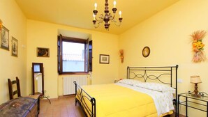 Une chambre à coucher comprenant un lit, une chaise, une commode, une fenêtre, un radiateur, un lustre et une table de chevet.