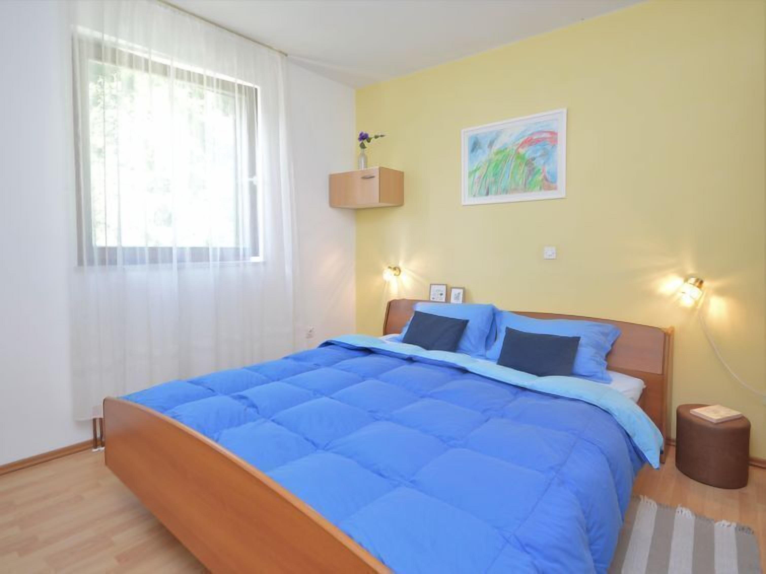A bedroom with a wooden bed, blue bedding, a window with white curtains, a wall-mounted shelf with a vase, and a painting.