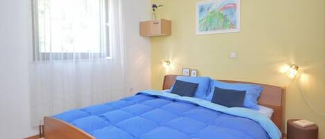 A bedroom with a wooden bed, blue bedding, a window with white curtains, a wall-mounted shelf with a vase, and a painting.