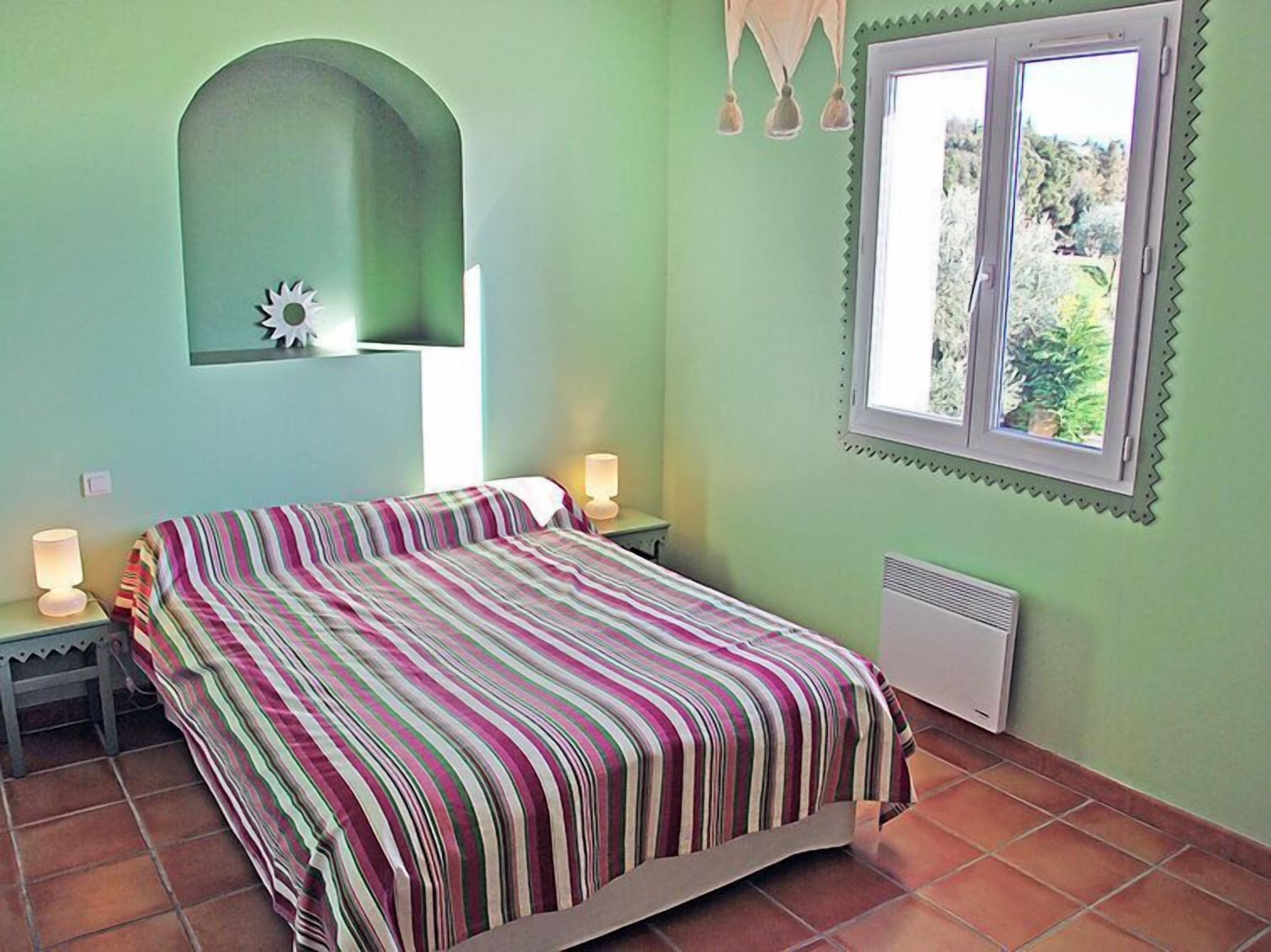 Un dormitorio con una cama, dos lámparas de noche, un espejo en la pared, una ventana con vistas a zonas verdes y un radiador.