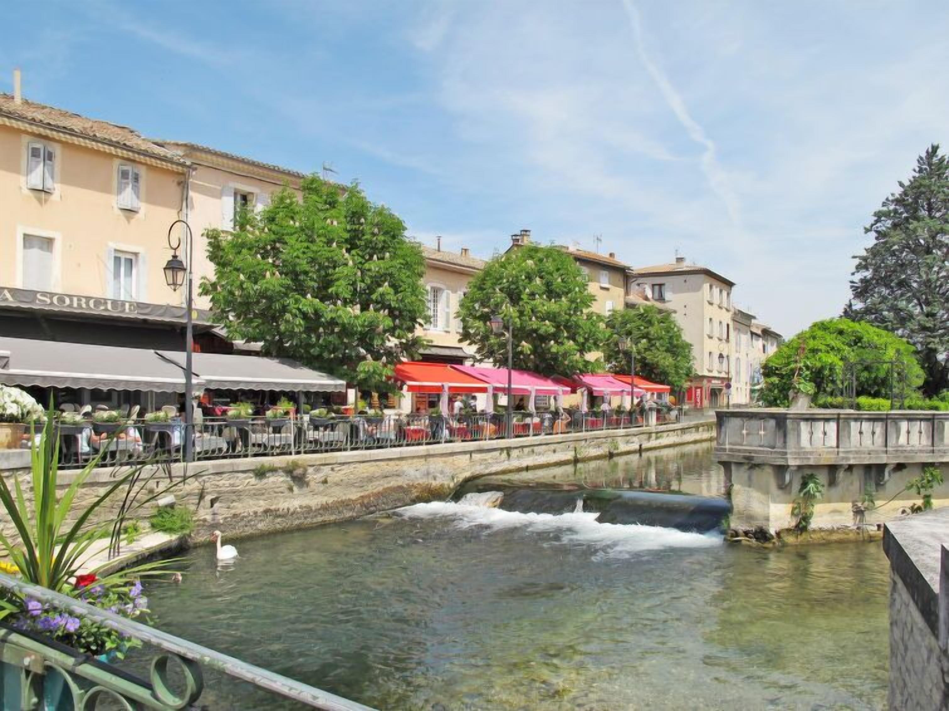 A canal with a small waterfall, surrounded by buildings with outdoor seating and greenery.