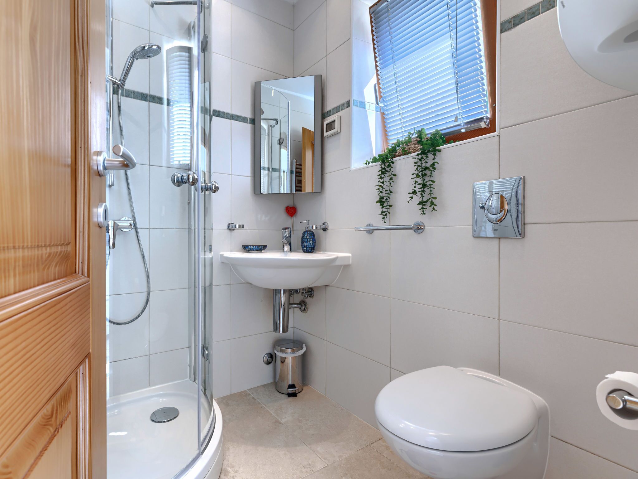 A modern bathroom with a glass-enclosed shower, a white sink, a toilet, and a window with blinds.