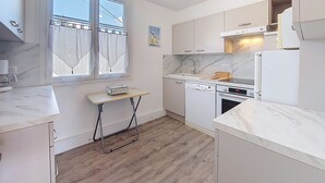 Moderni keittiö, jossa on valkoiset kaapit, sisäänrakennettu uuni ja astianpesukone. Siinä on ikkuna, jossa on verhot, ja pieni pöytä, jonka päällä on laatikko, jossa on merkintä ”SEASONS”.