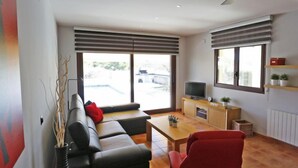 A modern living room with a sofa, a red chair, a television on a wooden cabinet, and large windows with blinds. - Perales by Interhome (L'Ampolla)