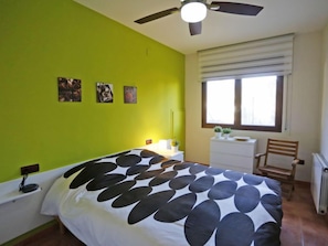 A bedroom with a bed, a ceiling fan, a window with blinds, and a small table with a chair.