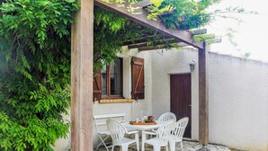 Une terrasse avec un sol en pierre, des chaises en plastique blanches, une table en bois et une pergola couverte de plantes grimpantes.