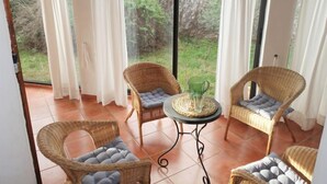 Une pièce avec des chaises en osier et une table, de grandes fenêtres avec des rideaux blancs, et une vue sur la verdure à l’extérieur.