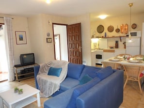 Sala de estar con un sofá azul, una butaca tapizada, una mesita con objetos decorativos, un televisor y una zona de cocina con una mesa de comedor para cuatro personas.