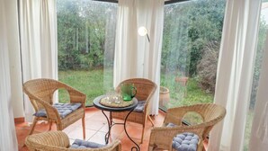 Une terrasse ensoleillée avec des fauteuils en osier et une table, de grandes fenêtres avec des rideaux et une vue sur la verdure.