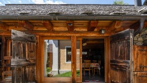 A wooden cabin with a stone entrance, a dining area visible through the glass door, and a small garden with flowers.