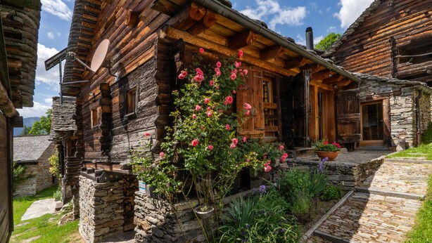 A rustic wooden house with a stone foundation, a wooden porch, and a garden with flowering plants.
