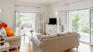 A spacious living room with a white sofa, a small table, and a TV on a cabinet. Large glass doors lead to an outdoor area with a pool and lounge chairs. - Villa Pescada by Interhome (Albufeira)
