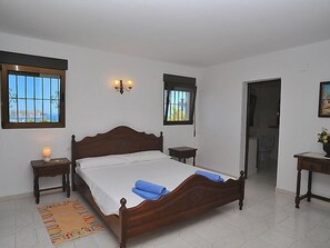 Une chambre à coucher avec un lit en bois, deux tables de chevet, une commode, une fenêtre donnant sur un beau paysage et une porte menant à une autre pièce.