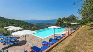 Un espace piscine agrémenté de chaises longues bleues et de parasols blancs, entouré d’une terrasse en pierre et offrant une vue sur un paysage montagneux.