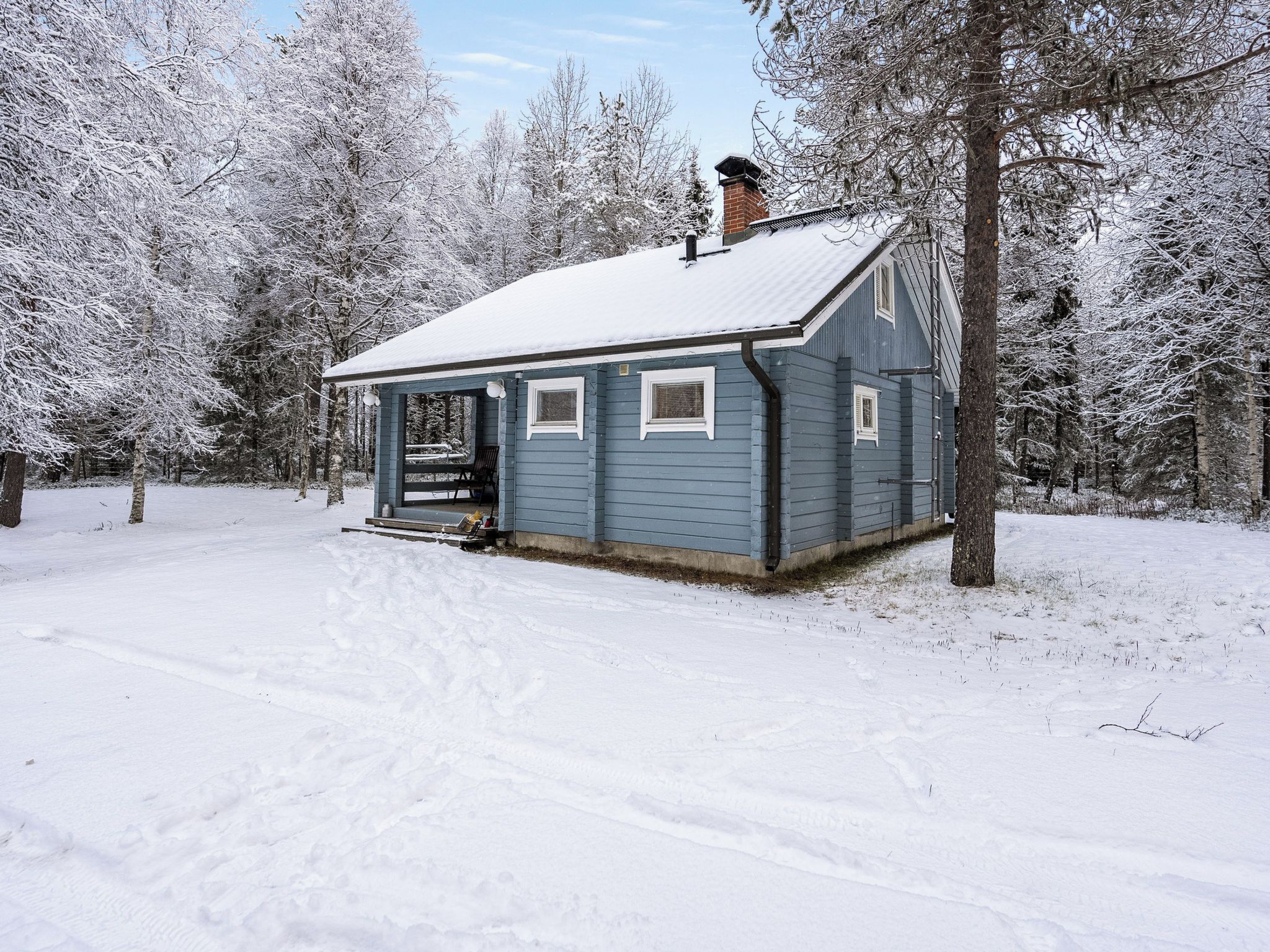 A small cabin with a porch and a chimney, surrounded by snow-covered trees.