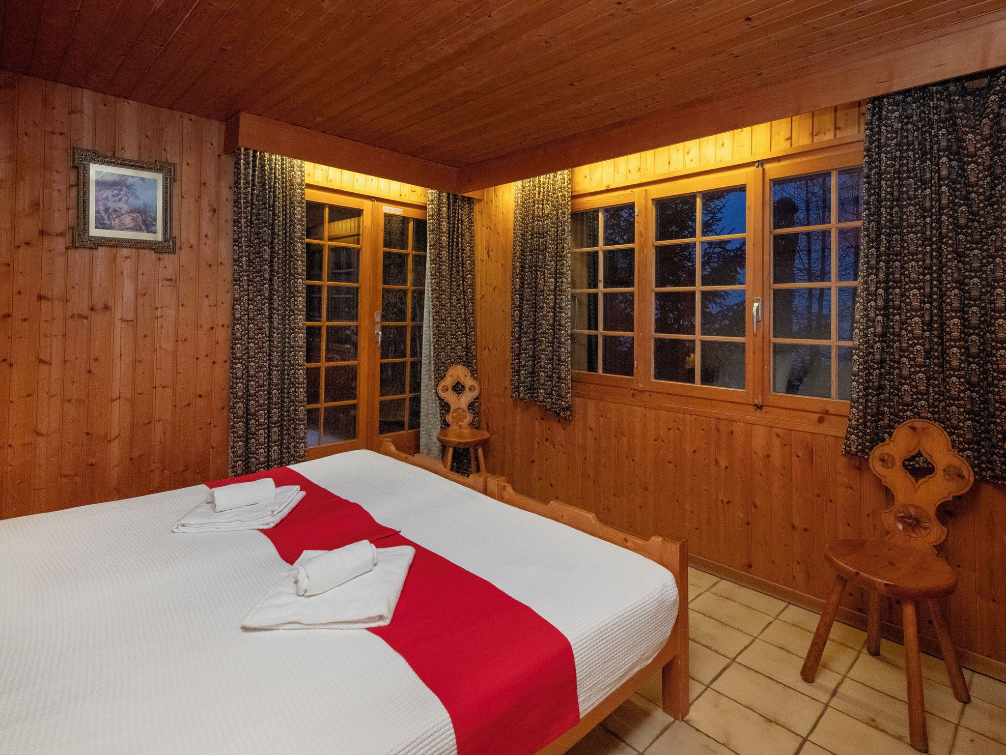 A wooden cabin room with a large bed, two windows with curtains, and a small wooden chair.