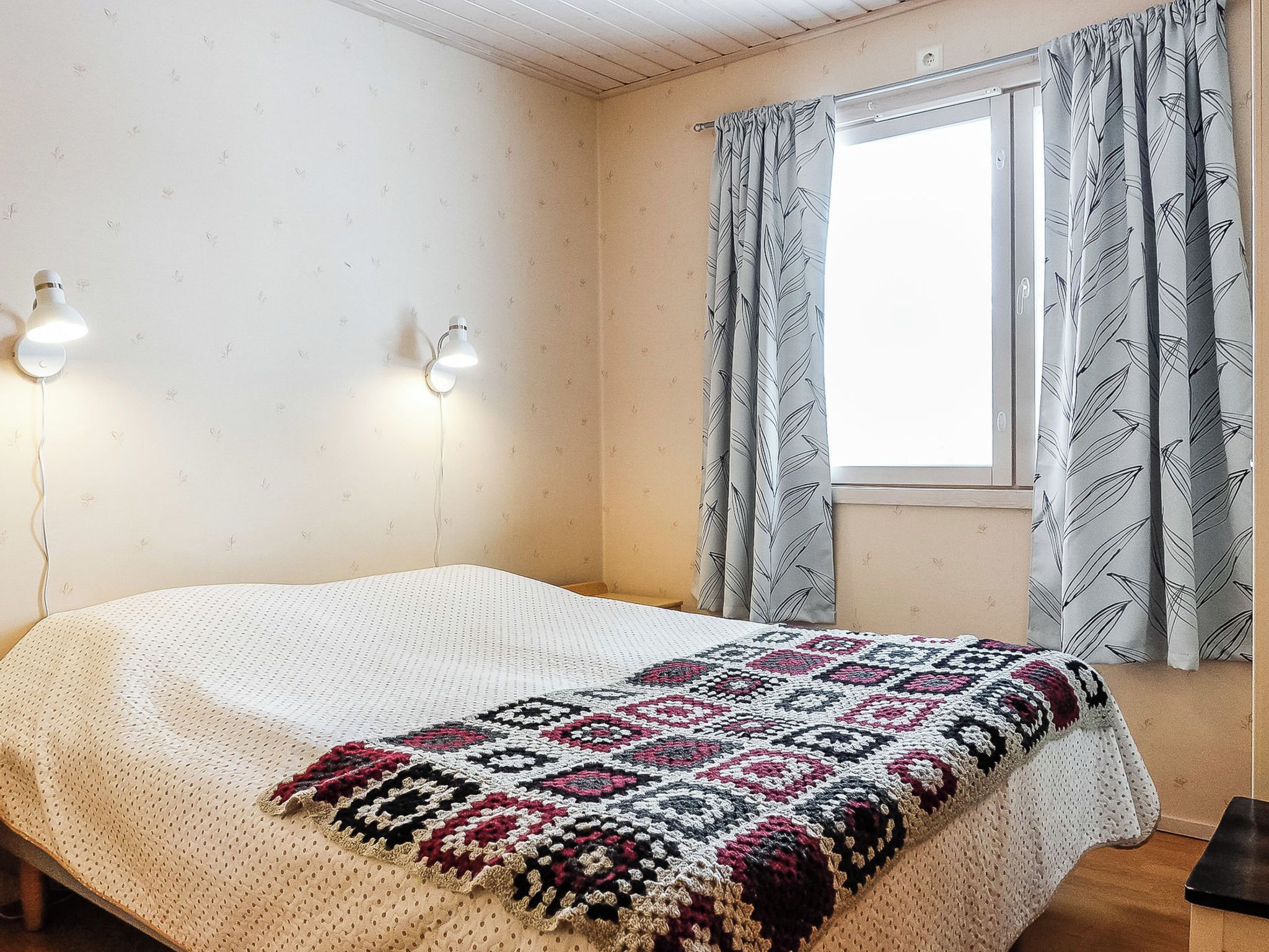 Une chambre à coucher avec un lit, deux lampes fixées au mur, une fenêtre avec des rideaux et une couverture à motifs.