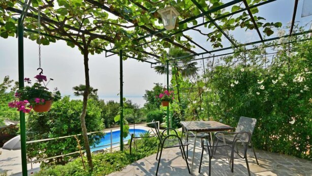 A covered patio with a table and chairs, a hanging flower pot, and a view of a pool and greenery. - Apartment Zvonko by Interhome (Lovran)