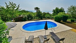 A swimming pool with a curved edge, surrounded by lounge chairs and umbrellas, with a view of the sea and greenery. - Apartment Zvonko by Interhome (Lovran)