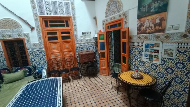 A traditional Moroccan interior with a patterned tile floor, a dining area with a table and chairs, and decorative elements like a wooden door and wall art. - Riad Drissia (Fes)