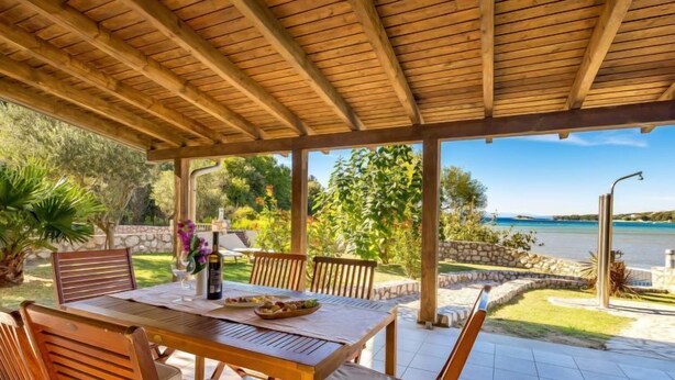 Een terras buiten met een houten tafel en stoelen, een fles wijn en een schaal fruit.