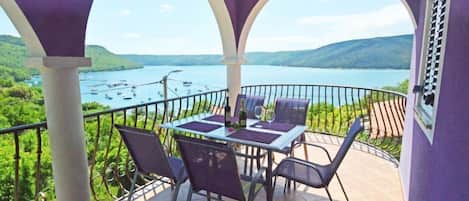 A balcony with a table and chairs, overlooking a lake with boats and hills.