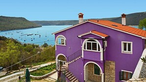 A two-story building with a red-tiled roof, white trim, and a purple exterior. It features a stone staircase leading to the upper floor and a view of a bay with boats. - Apartment Šverko by Interhome (Trget)
