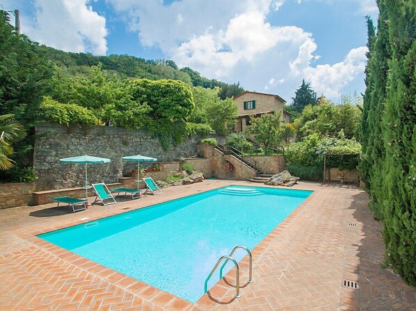 A swimming pool with lounge chairs and umbrellas, surrounded by greenery and a stone wall.